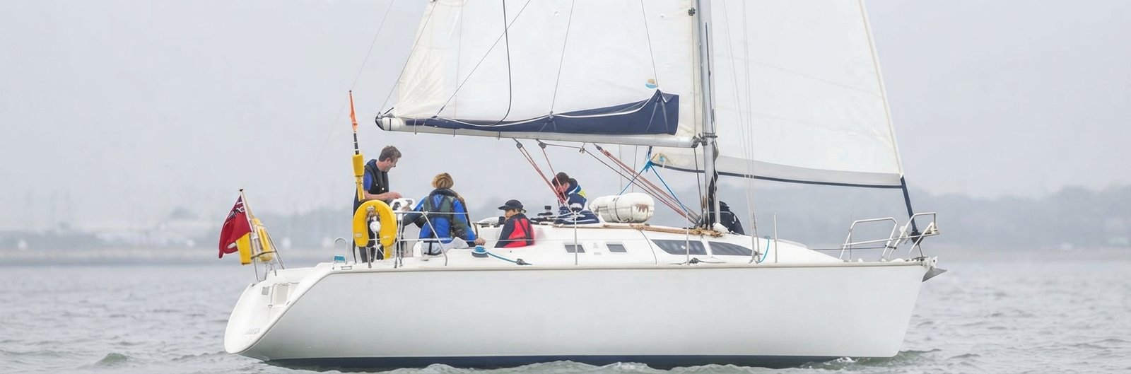 A side profile view of a white sailboat under sail on a calm, overcast day. Four crew members in lifejackets are on deck; one is at the helm, and others are working near the mast. A red ensign flag is flying from the stern. The background shows distant, hazy land.